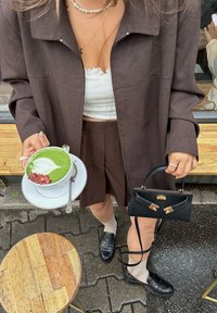 Veste marron oversize par-dessus un débardeur blanc raccourci, associée à une jupe plissée marron. Tenant une boisson verte dans une tasse blanche avec un motif de feuille. Sac à main noir et mocassins avec un détail texturé.