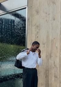 Mala mensageiro preta ao ombro, camisa branca de manga longa e calças escuras. Figura sorridente em frente a uma parede de vidro texturizado.