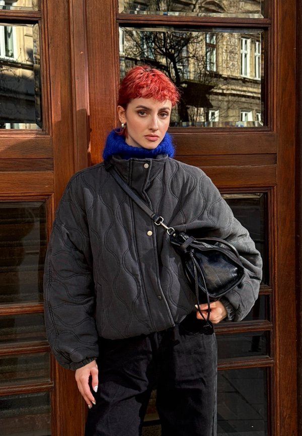 Person med kort rødt hår iført en sort quiltet jakke, blå højhalset trøje og sort taske, stående foran en trædør med glaspaneler.