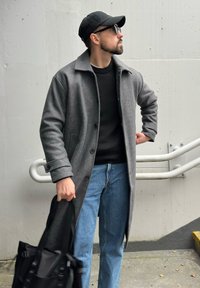 Manteau en laine gris avec col, associé à un pull noir et un jean bleu. Sac noir tenu à la main, sur fond de béton.