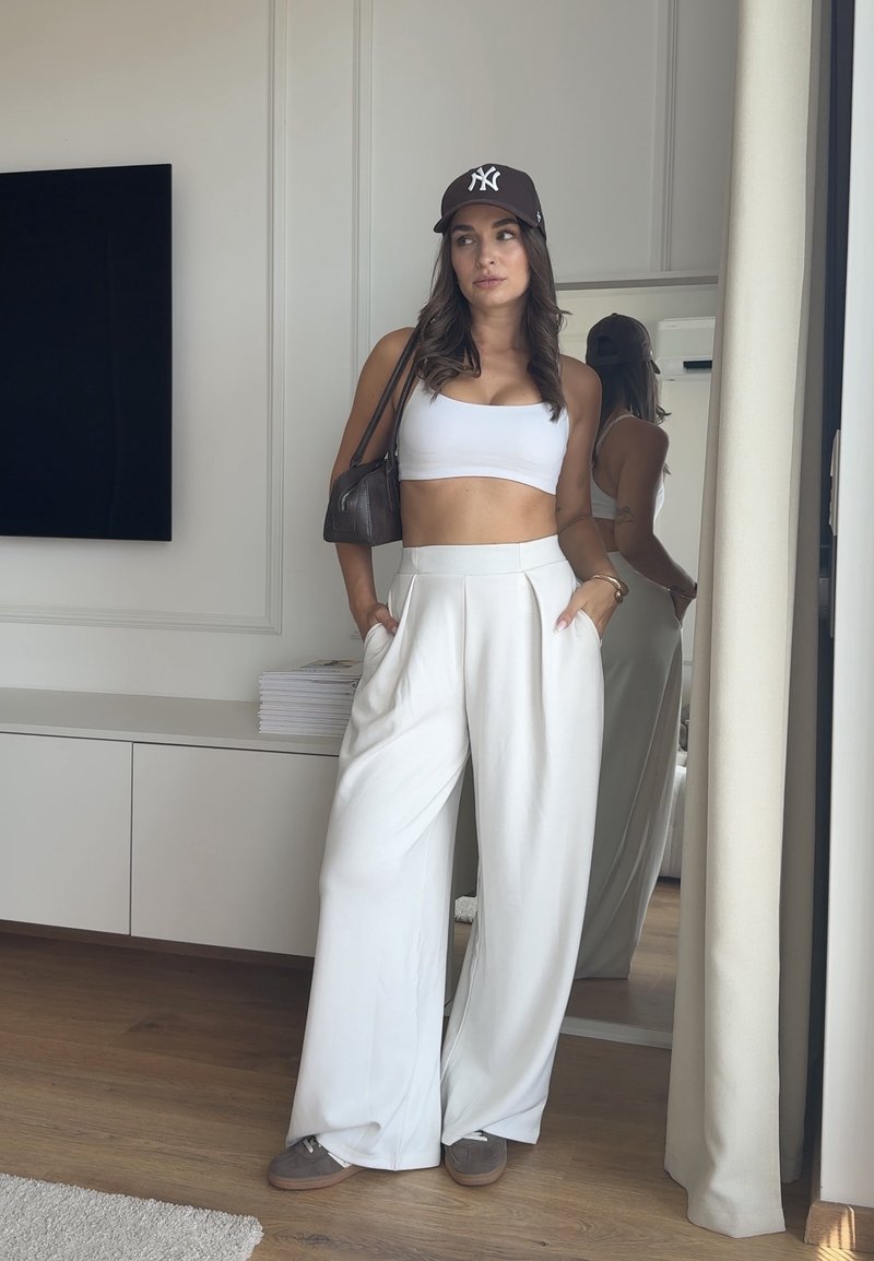 Γυναίκα με λευκό crop top, φαρδιά άσπρη παντελόνα, καφέ καπέλο και γκρι παπούτσια στέκεται μέσα σε χώρο με χέρια στις τσέπες και καφέ τσάντα στον ώμο.