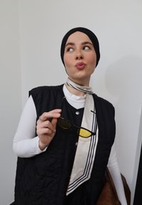 Femme portant un foulard noir et un gilet tient des lunettes de soleil à verres jaunes en regardant sur le côté, vêtue d'un haut blanc et d'un foulard rayé autour du cou.