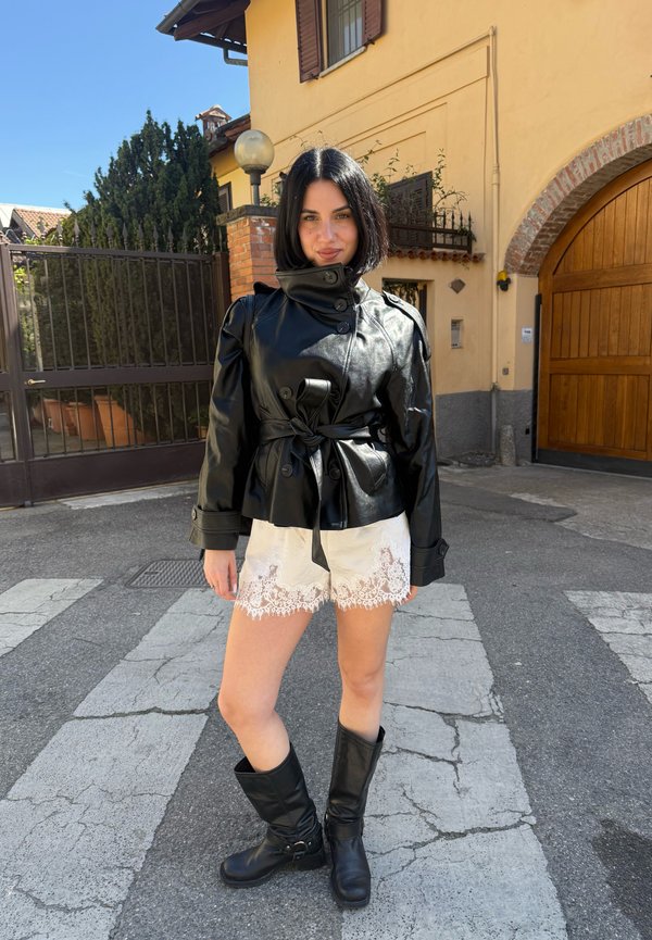 Žena s crnim bob kojim nosi crnu kožnu jaknu, bijele čipkane kratke hlače i crne čizme stoji na ulici s žutim zgradama iza sebe.