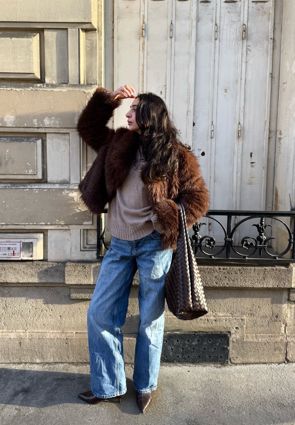 Femme en veste en fourrure marron, pull beige, jean large et chaussures pointues, debout sur un trottoir tenant un sac tote en rotin marron, se protégeant les yeux du soleil.
