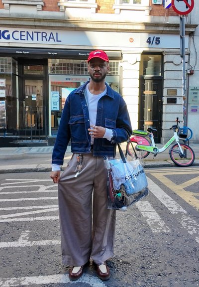 Chaqueta de denim, camiseta blanca, pantalones de pierna ancha beige, zapatos marrones. Bolsa transparente con contenido. De pie en la calle con bicicletas de fondo.