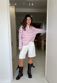 Camisa polo de mangas longas em rosa claro com finas riscas escuras, combinada com calções brancos e botas castanhas até ao joelho com fivelas.