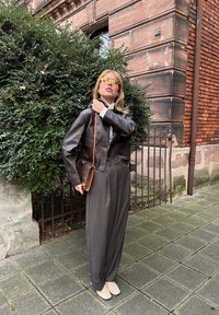 Braune Leder-Cropped-Jacke, graue schmal geschnittene Hose, beiges Oberteil, gelbe Sonnenbrille, trägt eine gemusterte Tasche, steht auf einem Steinpflaster.