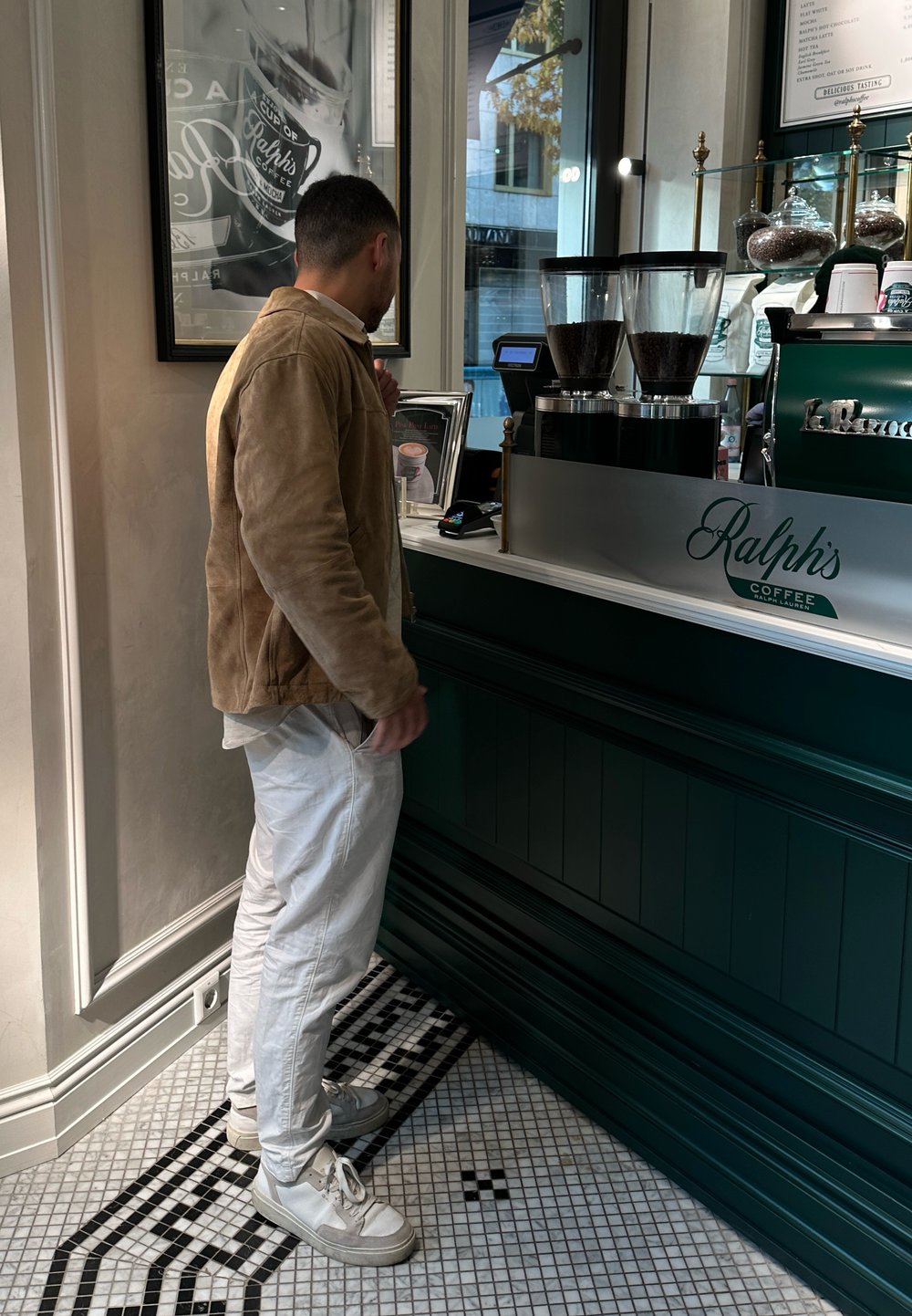 Comptoir à café avec un devant en bois vert foncé, des finitions en argent, des contenant transparents de grains de café et un sol en carreaux noirs et blancs.