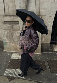 Veste en cuir marron, chemise rayée, pantalon large noir, chaussures noires à talons hauts, tenant une pochette ornée de fourrure, sous un parapluie noir.
