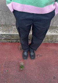 Μαύρα cargo παντελόνια με πλαϊνές τσέπες, συνδυασμένα με μαύρα παπούτσια και ένα πλεκτό πουλόβερ σε πράσινο και ροζ χρώμα. Το έδαφος είναι κόκκινο με εμφανή υφή.