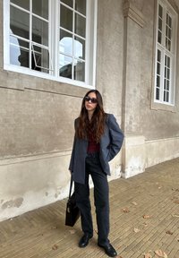 Blazer oversized cinza, top bordô, jeans pretos e sapatos pretos. Segurando uma bolsa preta, de pé sobre um calçamento de pedras com folhas caídas.