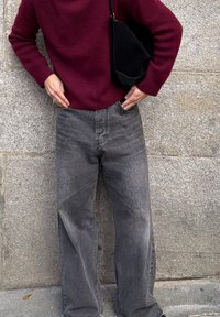 Jean gris à jambes larges avec une légère texture délavée, associé à un pull bordeaux. La personne tient un sac à épaule noir et pose une main dans la poche.