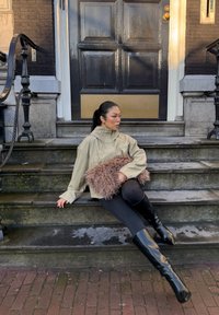Femme en veste beige et bottes noires assise sur des marches en pierre, tenant un sac brun en fourrure devant une porte en bois sombre.