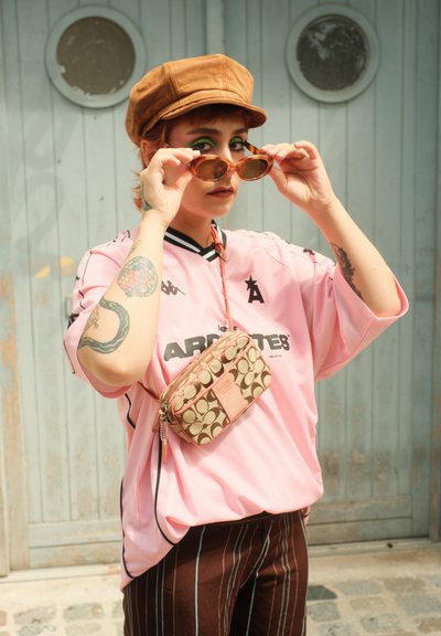 Jersey atlético rosa con acentos negros, pantalones a rayas marrones, gorra plana color beige y un pequeño bolso cruzado con un patrón marrón; gafas de sol en la mano.