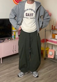 Szara bluza rozpinana na zamek, na białej koszulce z nadrukiem, w zestawieniu z luźnymi, ciemnozielonymi spodniami i srebrnymi tenisówkami. Wnętrze z kolorową dekoracją.