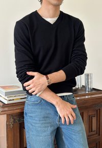 Mand iført sort V-hals sweater, blå jeans og et brun læderur, stående med armene krydsede foran et træskab.