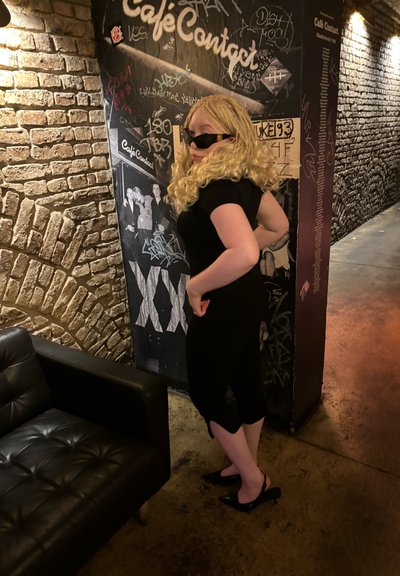 Ropa negra, peluca rubia rizada, gafas de sol y tacones. De pie contra una pared de ladrillo texturizada con grafitis. Sofá de cuero negro cerca.