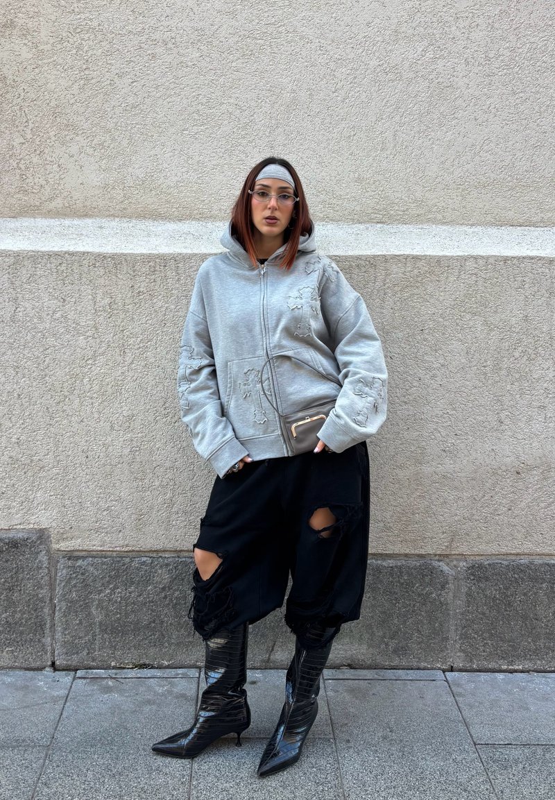 Young person in gray hoodie, black ripped pants, black pointed boots, and headband stands against textured beige wall.