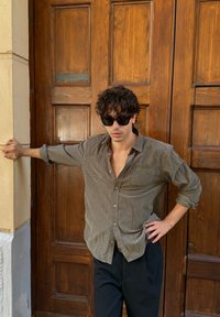 Giovane con capelli ricci e occhiali da sole, che indossa una camicia grigia arrotolata e pantaloni neri, appoggiato a una porta e a un muro in legno.