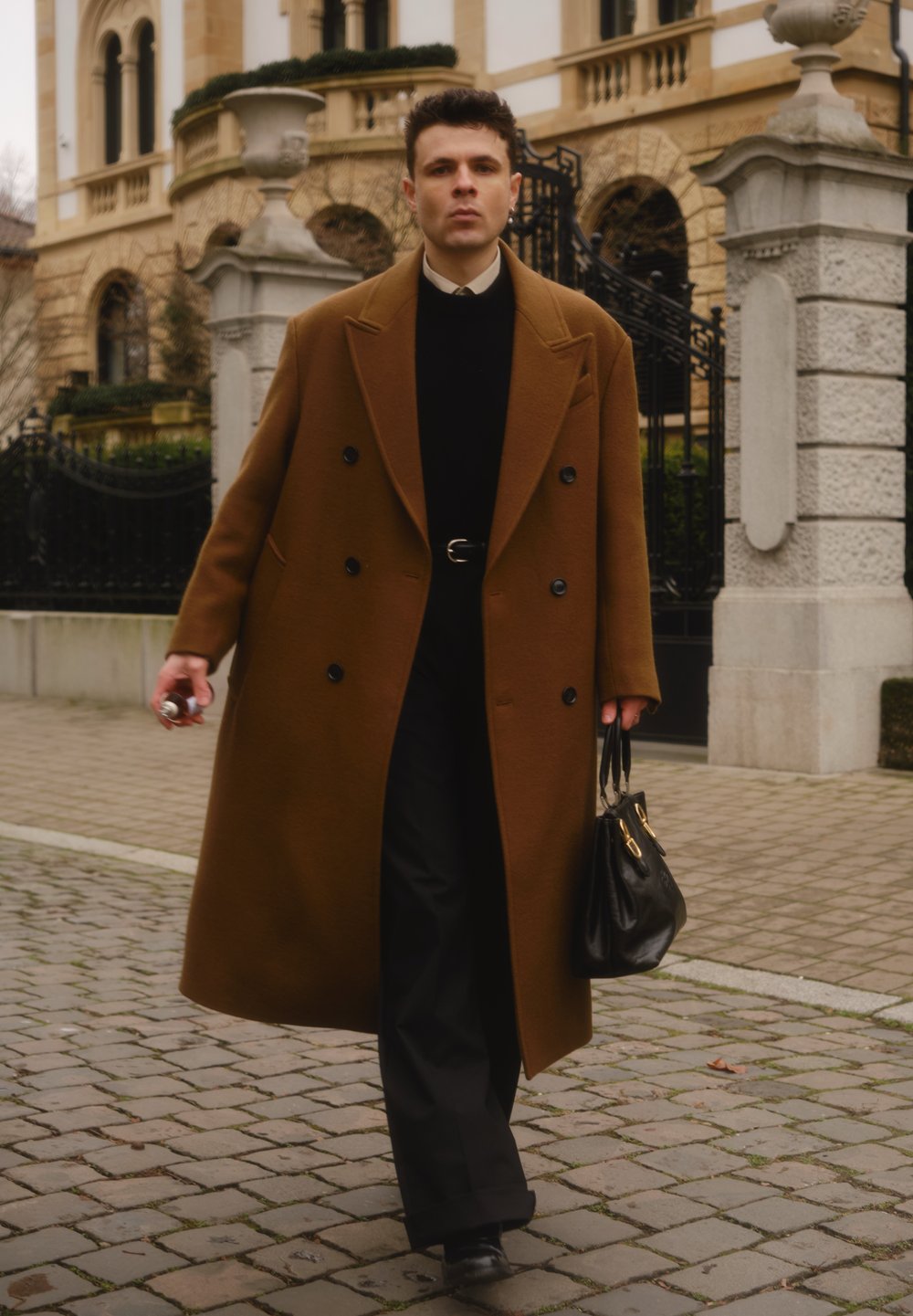 Manteau marron croisé, pull noir, pantalon noir, tenant un petit sac à main noir. Sol pavé et bâtiment historique en arrière-plan.