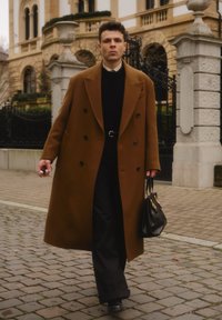 Manteau marron croisé, pull noir, pantalon noir, tenant un petit sac à main noir. Sol pavé et bâtiment historique en arrière-plan.