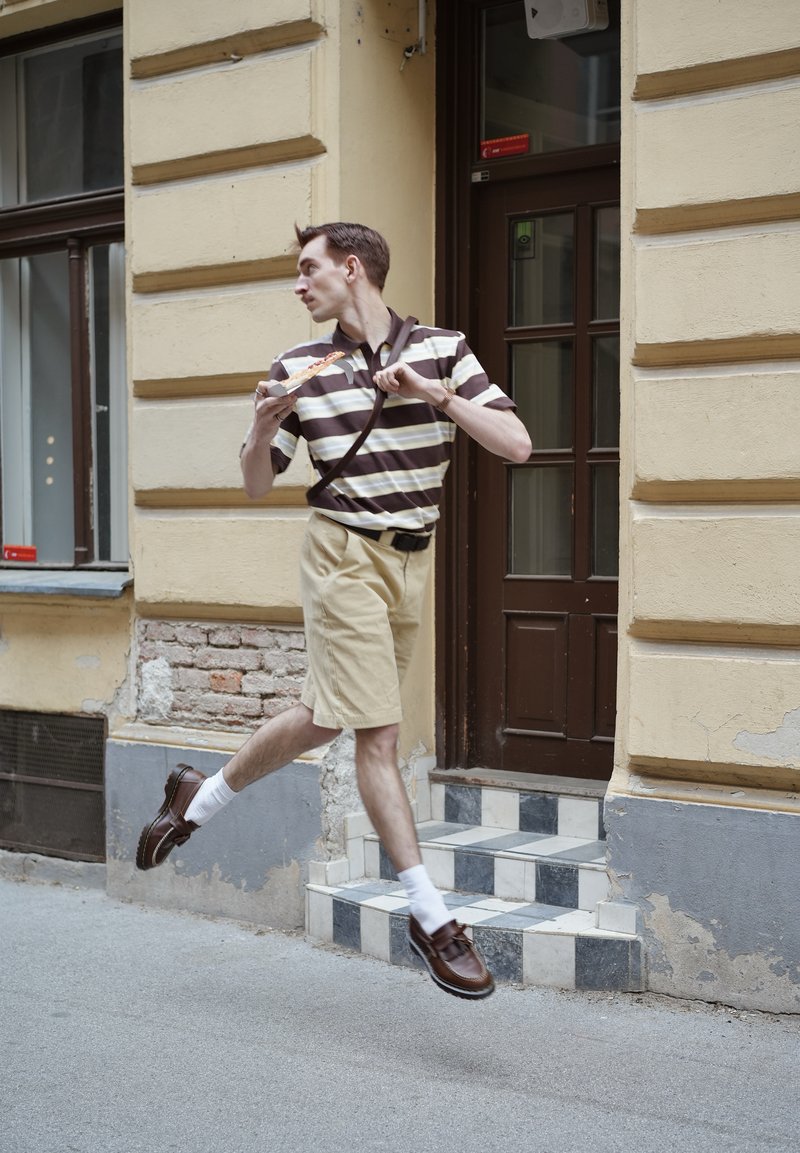 Homme en chemise rayée et short en plein saut près de l'entrée du bâtiment, tenant une part de pizza et regardant sur le côté.