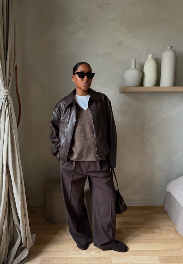 Woman wearing dark sunglasses, brown leather jacket, brown knitted vest, loose brown trousers, and holding a small brown bag in a minimalistic room setting.
