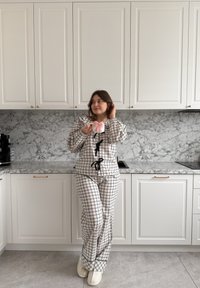 Femme en pyjama à carreaux tenant une tasse blanche avec un nœud rose, debout dans une cuisine moderne avec des armoires blanches et un plan de travail en marbre.