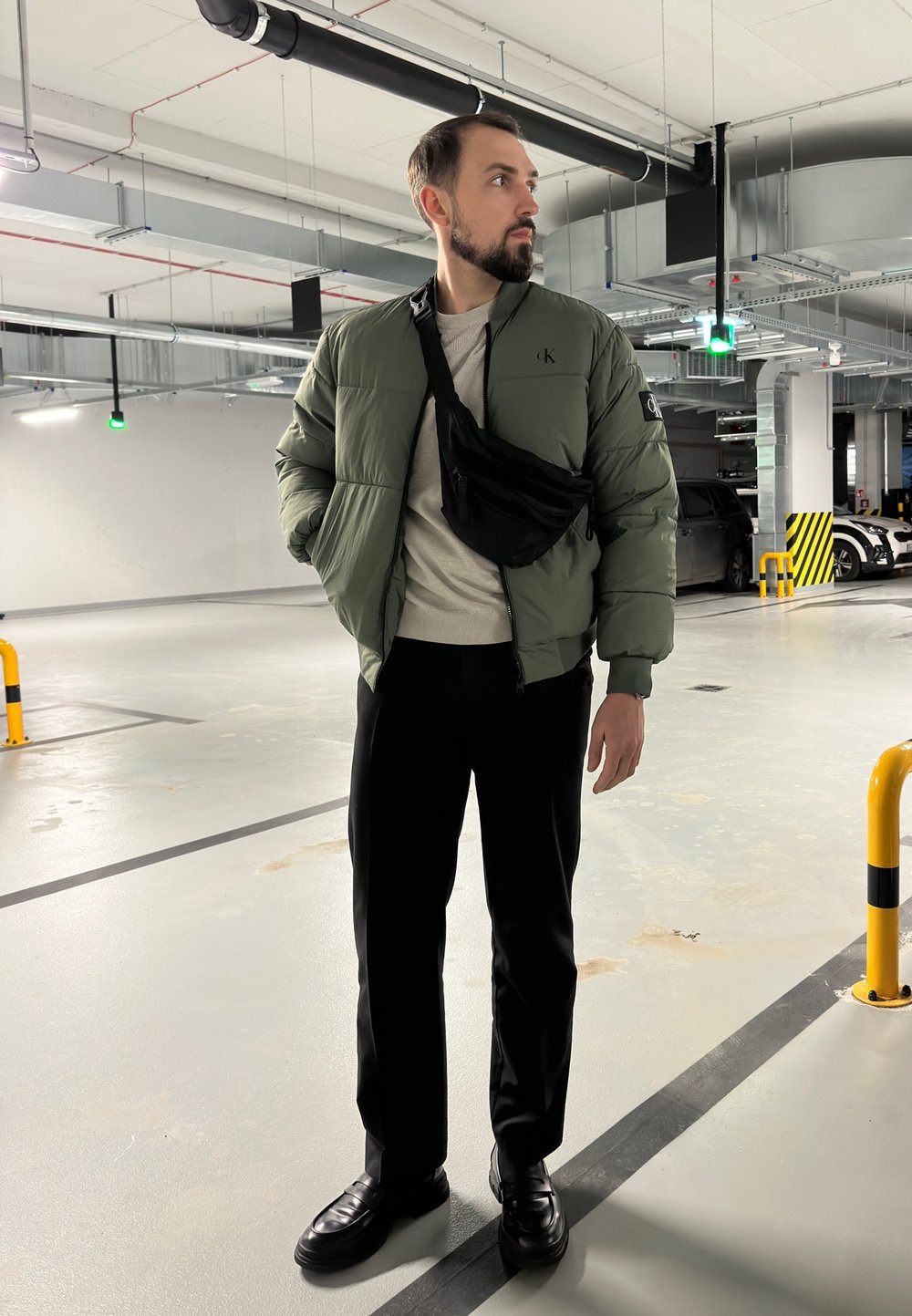 Daunejacke in Olivgrün, schwarze Umhängetasche, beiger Pullover, schwarze Hose und schwarze Slipper, in einer Parkgarage.