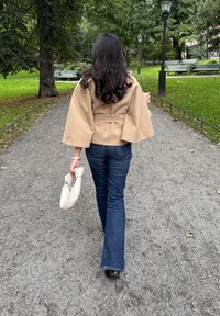 Kvinna med långt mörkt hår, iklädd beige jacka och blå jeans, som går på en grusväg i en grön park och håller en vit handväska.