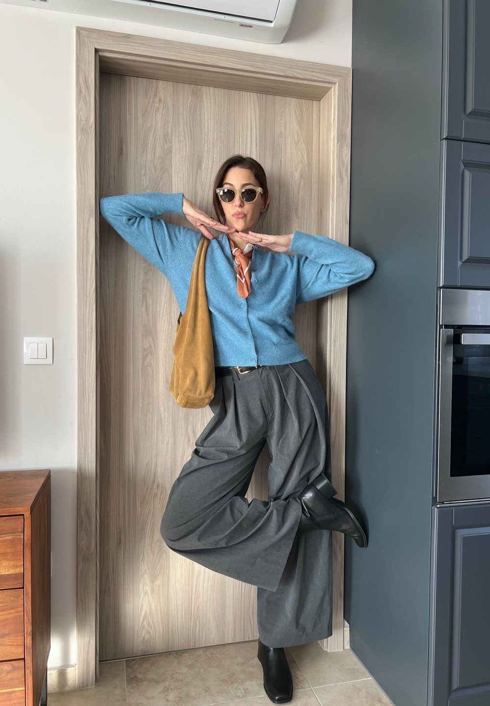 Ανοιχτό μπλε καζάκα πάνω από ένα λευκό πουκάμισο, συνδυασμένο με γκρι φαρδύ παντελόνι και μαύρες μπότα ankle. Κρατάει μια ταμπα σουέτ τσάντα. Στρογγυλά γυαλιά ηλίου.