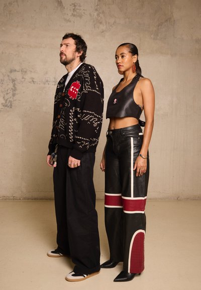 Hombre con cárdigan negro con estampado y pantalones holgados está junto a una mujer con top corto negro y pantalones de cuero negros con franjas rojas y blancas.