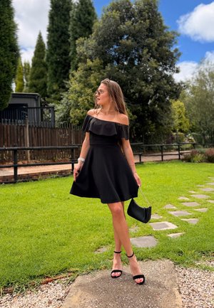 Mujer con vestido negro de hombros descubiertos y tacones, sosteniendo un bolso negro, de pie en un camino de piedra en un jardín verde con árboles y cielo azul.