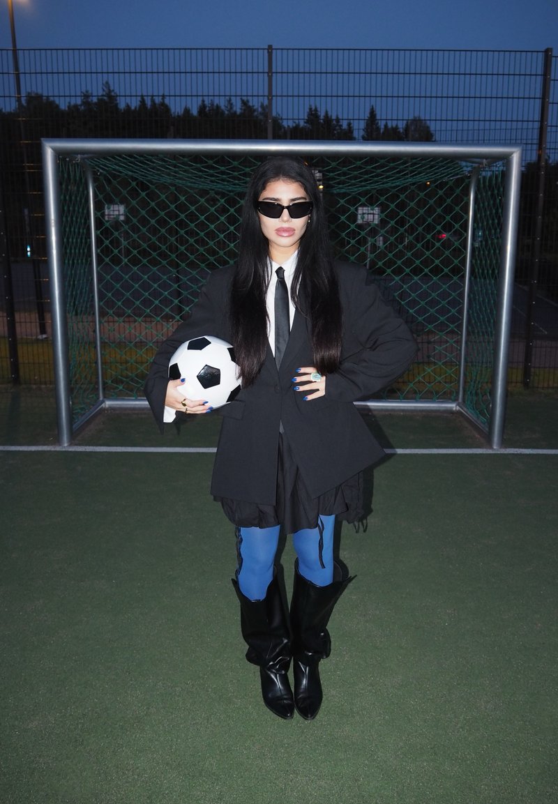 Black oversized blazer, white shirt, black tie, blue leggings, and knee-high black boots. Holding a black-and-white football in front of a goal.