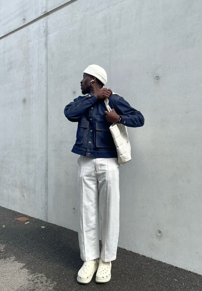Chaqueta de denim con bolsillos frontales, gorro blanco, bolso de tela color crema, pantalones anchos blancos y zuecos beige. Fondo de concreto.