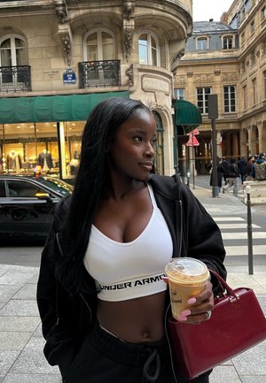 Mujer joven en sujetador deportivo blanco y chaqueta negra sostiene un café helado y un bolso rojo, de pie en una calle de París con edificios históricos.