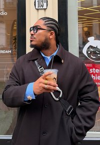 Abrigo negro con cuello, camisa azul claro. Sosteniendo un vaso transparente con una pajilla negra. Pelo trenzado y gafas de sol; un mosquetón plateado en la correa.