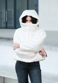 Veste matelassée blanche avec un col montant et une capuche surdimensionnée, présentant une texture lisse. Associée à un jean en denim foncé, tenant des lunettes de soleil.