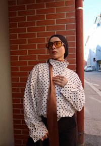 Persona vestida con una camisa blanca de lunares, un pañuelo oscuro en la cabeza, grandes gafas de sol y una corbata marrón, se encuentra de pie frente a una pared de ladrillos rojos al aire libre.