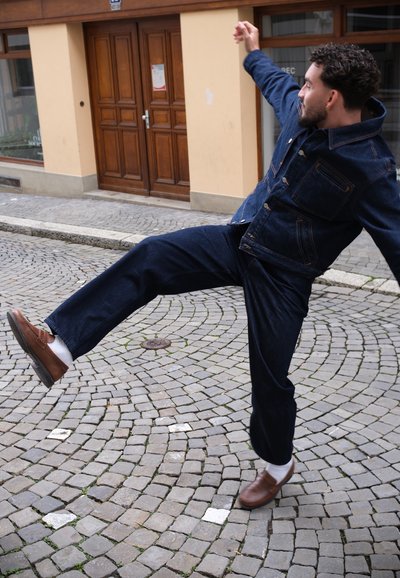 Chaqueta y pantalones de mezclilla en azul oscuro, con bolsillos y botones. Zapatos marrones con calcetines blancos, de pie en una calle de adoquines.