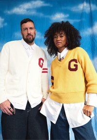 Two individuals are dressed in garments: one wears a cream cardigan adorned with a red letter and stripes, while the other sports a yellow sweater with brown accents, both layered over white shirts. Denim bottoms are also visible.