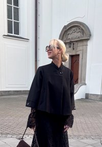 Czarna krótka kurtka z wysokim kołnierzem i zapięciem na guziki z przodu, nałożona na czarną koronkową sukienk�ę. Widać duże ciemne okulary przeciwsłoneczne oraz torebkę.