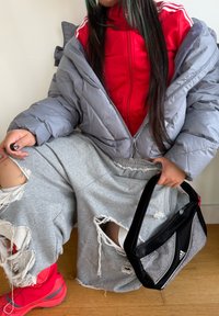 Harmaa toppatakki, punainen vetoketjullinen takki, repeytynyt harmaa verkkarit, punaiset kengät ja musta laukku hopeisilla yksityiskohdilla. Taustalla puiset lattialaudat.