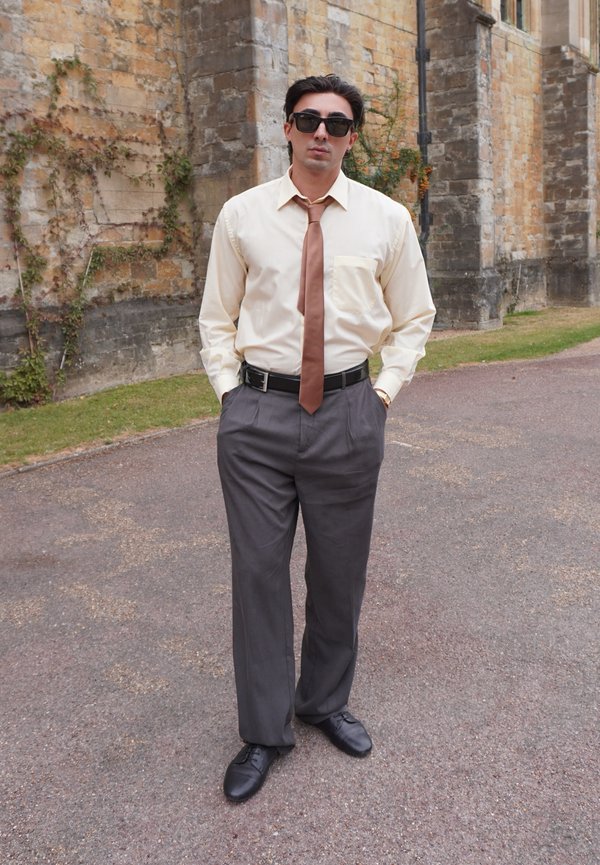 Homme portant des lunettes de soleil, une chemise crème, une cravate marron et un pantalon gris, debout sur un chemin pavé à côté d'un bâtiment en pierre avec des vignes.