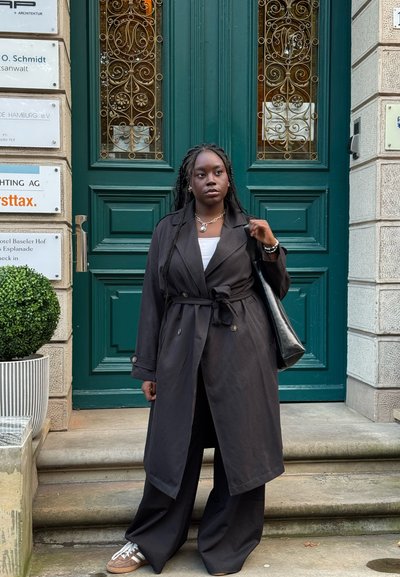 Abrigo negro sobre una blusa blanca, pantalones anchos negros y zapatillas. Bolso negro y cabello trenzado. Puerta verde con detalles decorativos en el fondo.