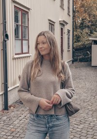 Pullover de malha bege com mangas largas, combinado com jeans azul claro. Uma pequena bolsa de ombro e um anel são visíveis. Fundo de calçada.