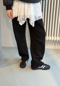 Fekete, laza szabású melegítőnadrágok, réteges fehér csipkés ruhadarabbal és fekete sportcipőkkel, amelyeken három fehér csík van.