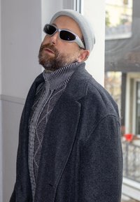 Manteau gris par-dessus un pull à motifs à col montant, complété par un bonnet gris clair et des lunettes de soleil blanches avec des verres foncés.