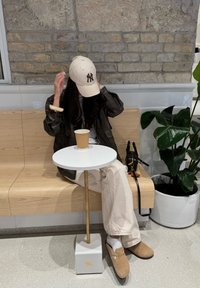 Hnědá bomber bunda, bílé tričko, krémové kalhoty, světle hnědé semišové boty. Kulatý bílý stůl se zlatou základnou; papírový kelímek; poblíž je květináč s rostlinou.