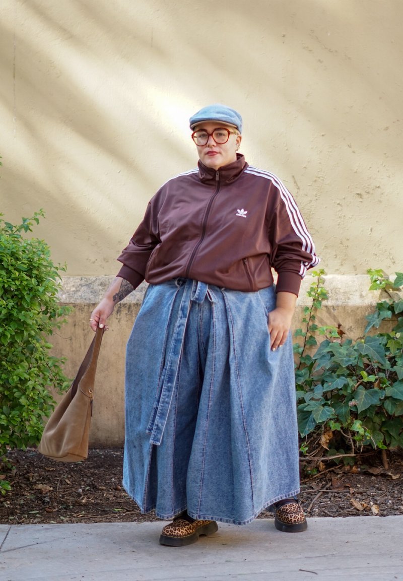 Braune Zip-Jacke mit weißen Streifen, blaue weite Denim-Hosen, Schuhe mit Leopardenmuster und eine graue Mütze, die eine braune Tasche hält. Grüne Blattpflanzen im Hintergrund.